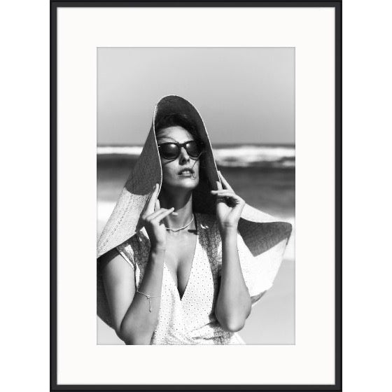 Black and white photograph of a woman in sunglasses and an oversized straw hat by the sea