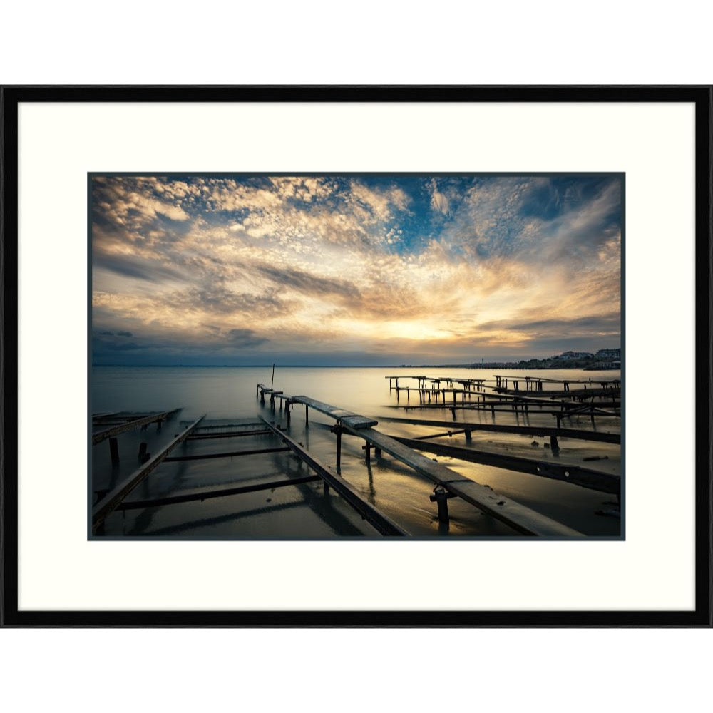 Photograph of harbor pier pilings under a dramatic golden dusk sky, black frame with white mat