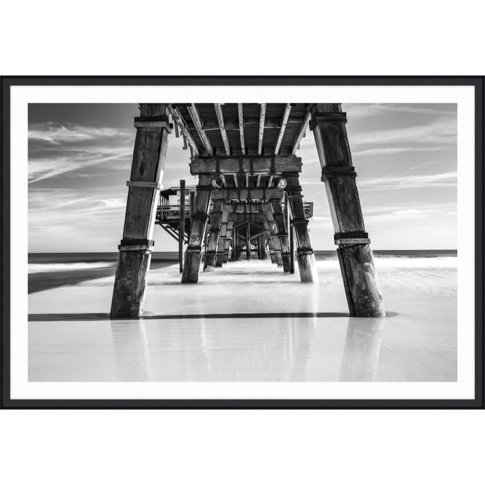 Black and white photograph from beneath a long wooden pier looking out to sea