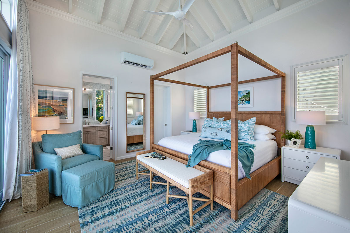 A bright, modern bedroom featuring a tall wooden four-poster bed with white bedding and blue accents, a matching blue armchair, a patterned blue and white rug, and white walls with exposed ceiling beams.