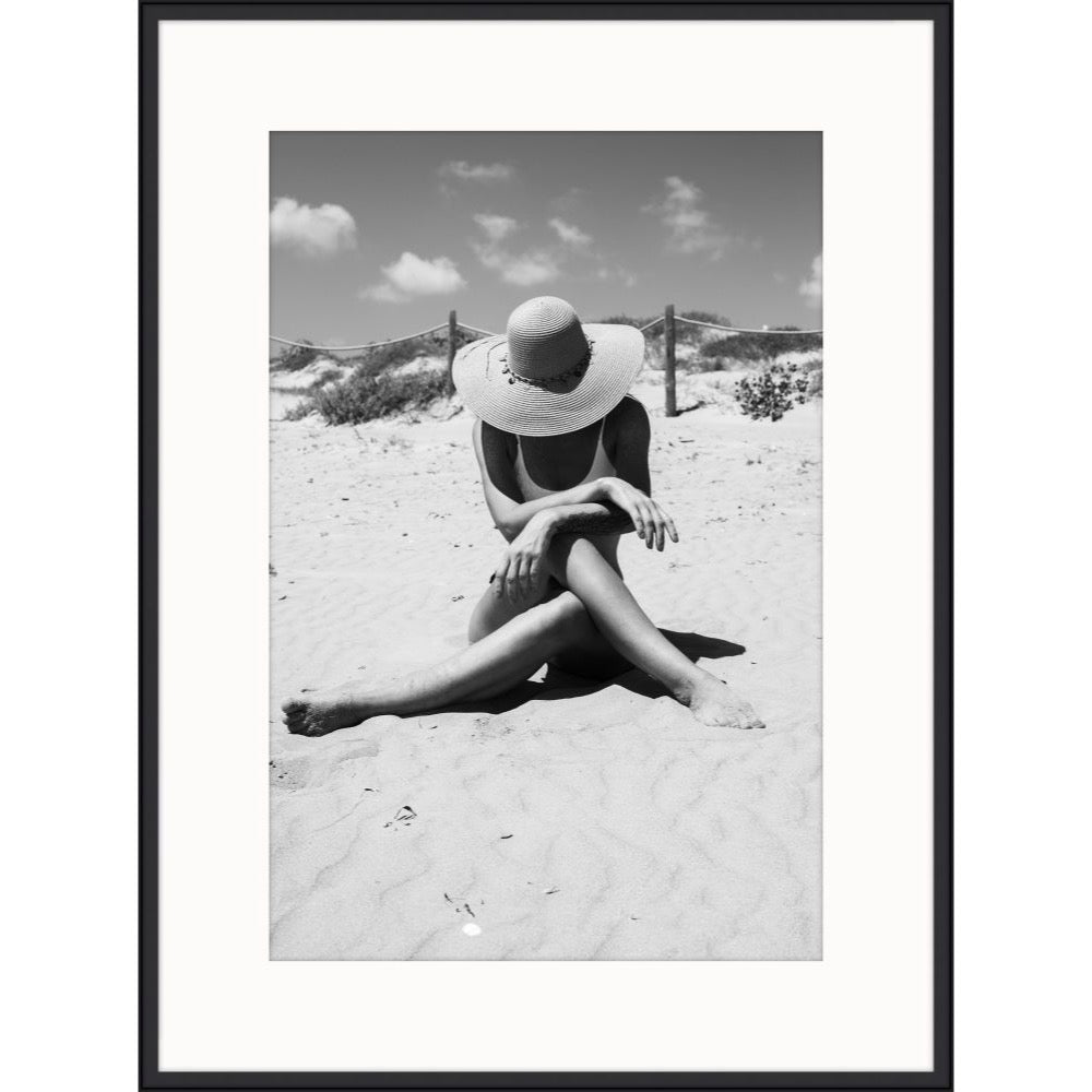 Black and white photograph of a woman in a wide sun hat sitting on rippled sand
