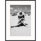 Black and white photograph of a woman in a wide sun hat sitting on rippled sand