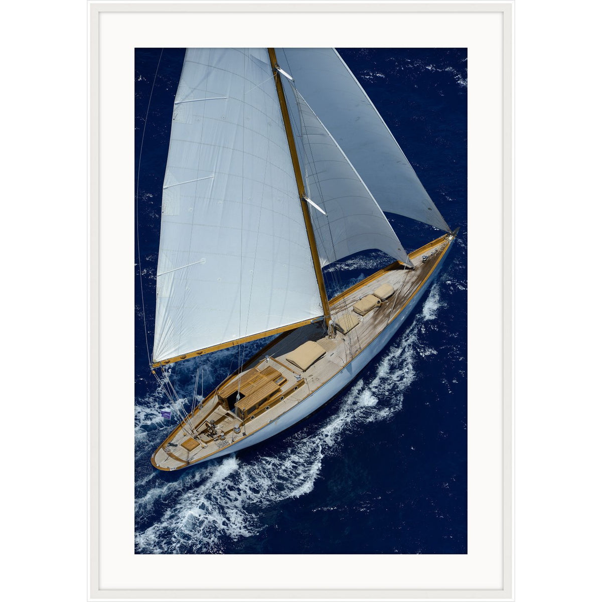 Aerial photograph of a classic wooden sailboat with white sails on deep blue water, framed with a white mat