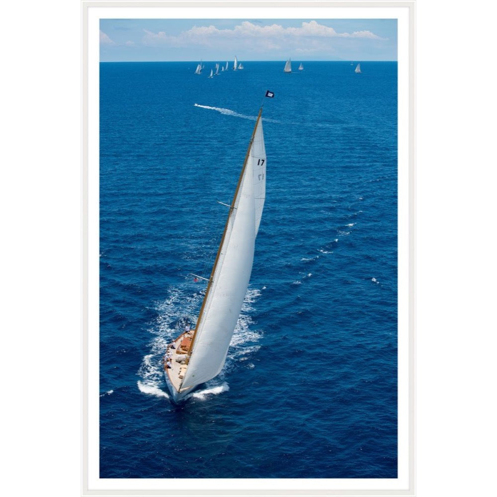 Aerial photograph of a white-sailed sloop leading a regatta on deep blue water