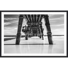 Black and white photograph from beneath a long wooden pier looking out to sea
