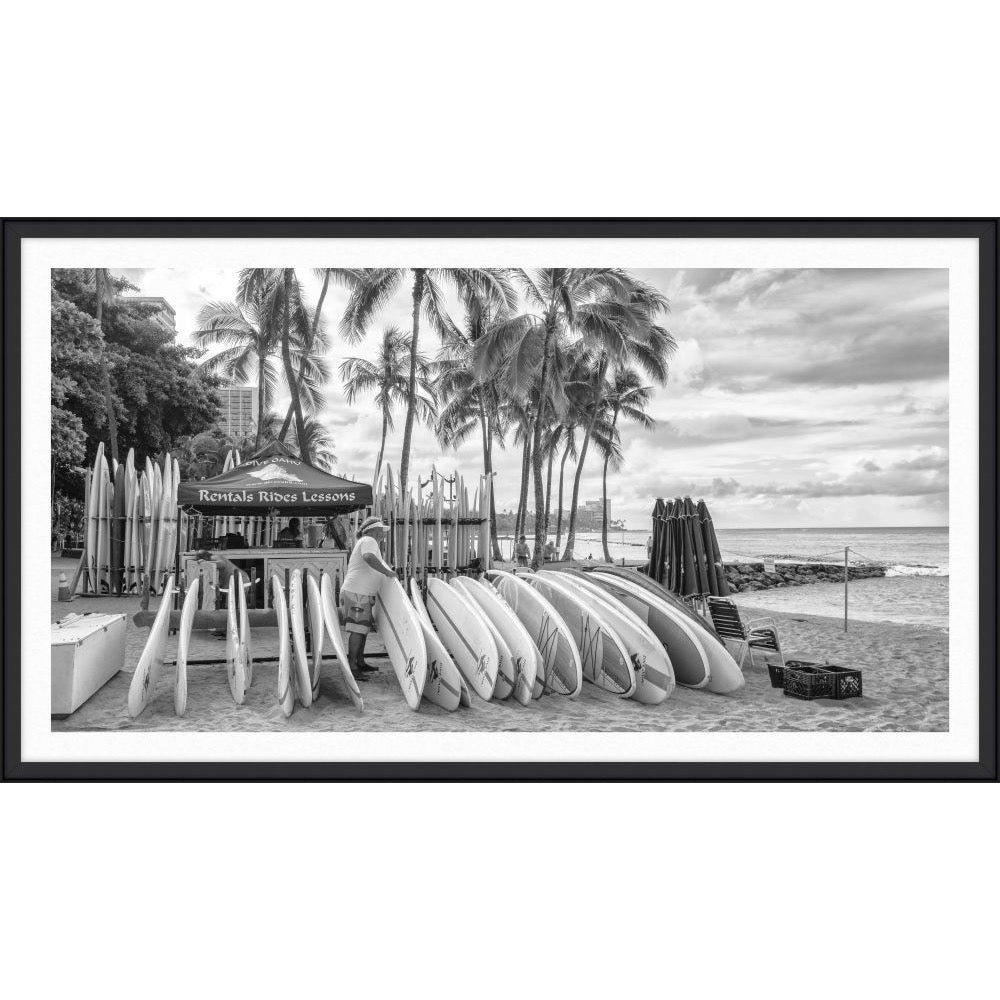 Black and white photograph of a surfboard rental stand with a row of surfboards under palm trees