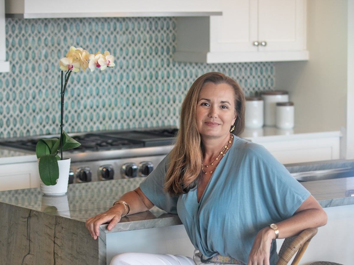 A woman with blonde hair wearing a light blue top and white pants sits at a kitchen island, with a patterned teal backsplash and white cabinets in the background.