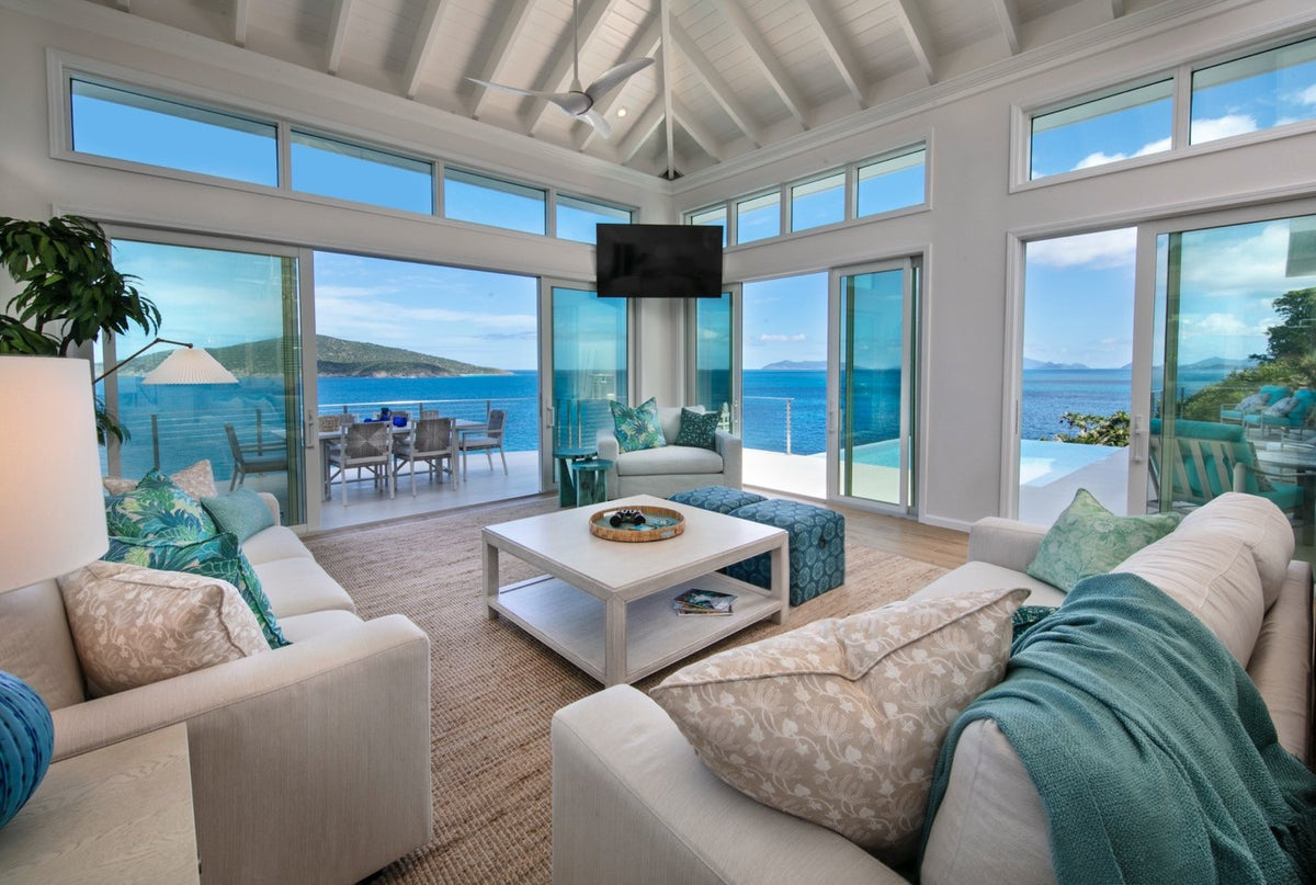Bright modern living room with white sofas, patterned pillows, a white coffee table, and large windows overlooking the ocean and patio.