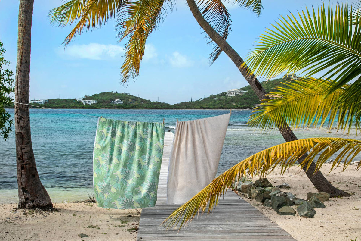 A tropical beach scene with two towels hanging on a string line outdoors between palm trees. One towel has a green palm leaf pattern, and the other is solid off-white.