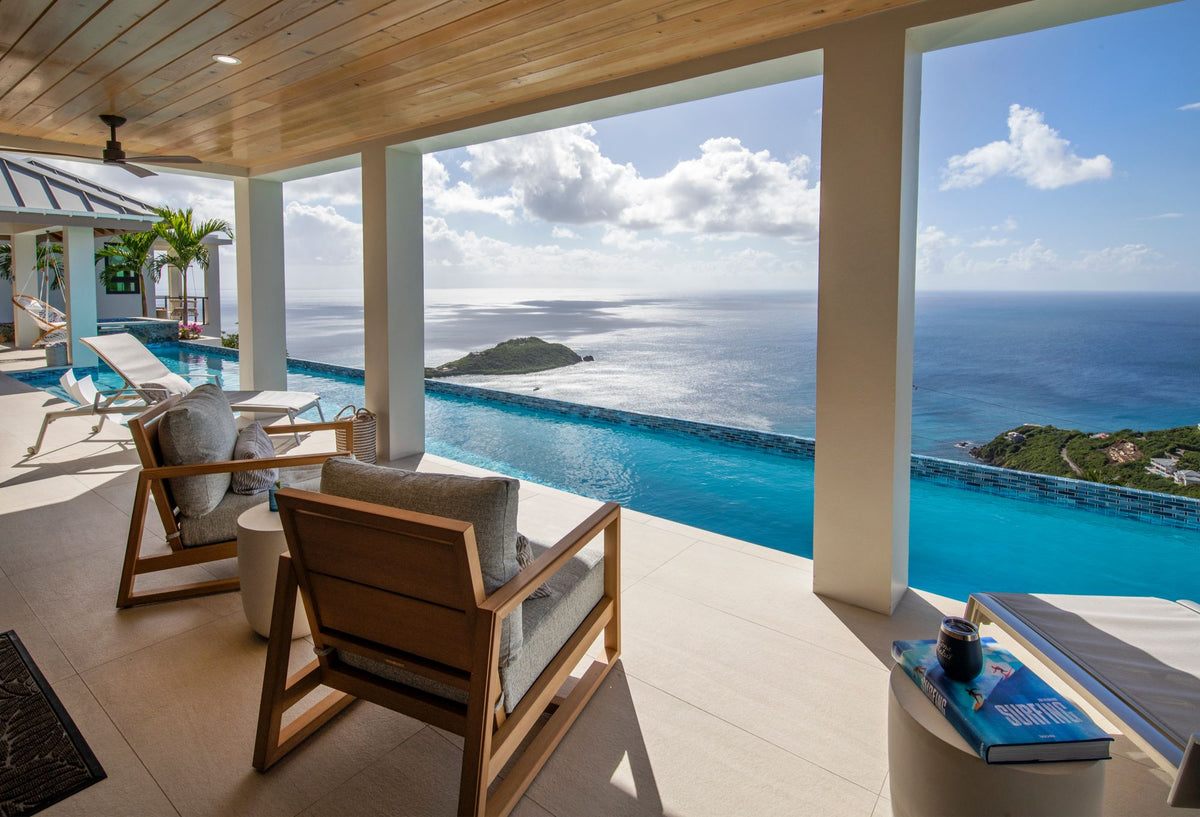 Modern outdoor patio seating area with two wooden armchairs and a round side table, overlooking an infinity pool and a scenic ocean view with clouds and a small island.