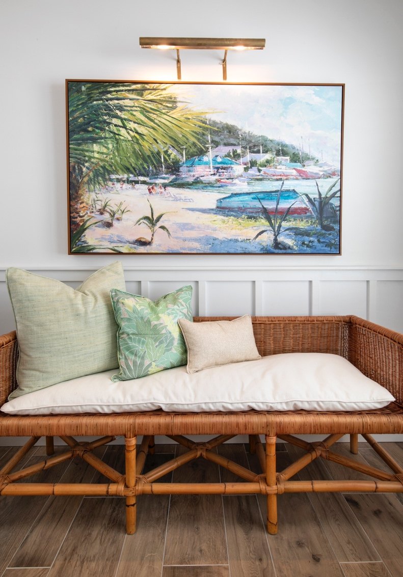 A framed tropical beach painting hangs on a white wall above a woven rattan daybed with cream cushions and green patterned pillows. A wall-mounted light fixture is visible above the artwork.