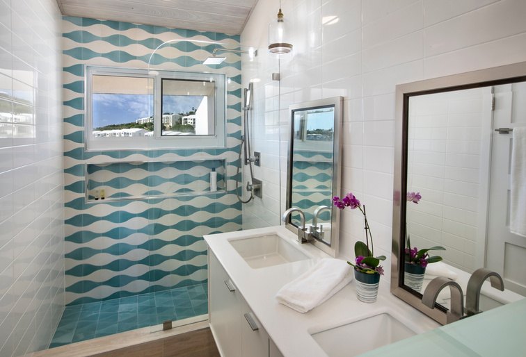 A modern bathroom interior with a walk-in shower featuring teal and white wavy patterned tile, white vanity cabinets, double sinks, mirrors, and small potted purple flowers.