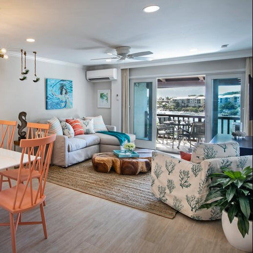 A bright, modern open-concept living room and dining area with light wood floors, a white dining table with coral chairs, a beige sectional sofa, a patterned armchair, and large glass doors leading to a balcony.