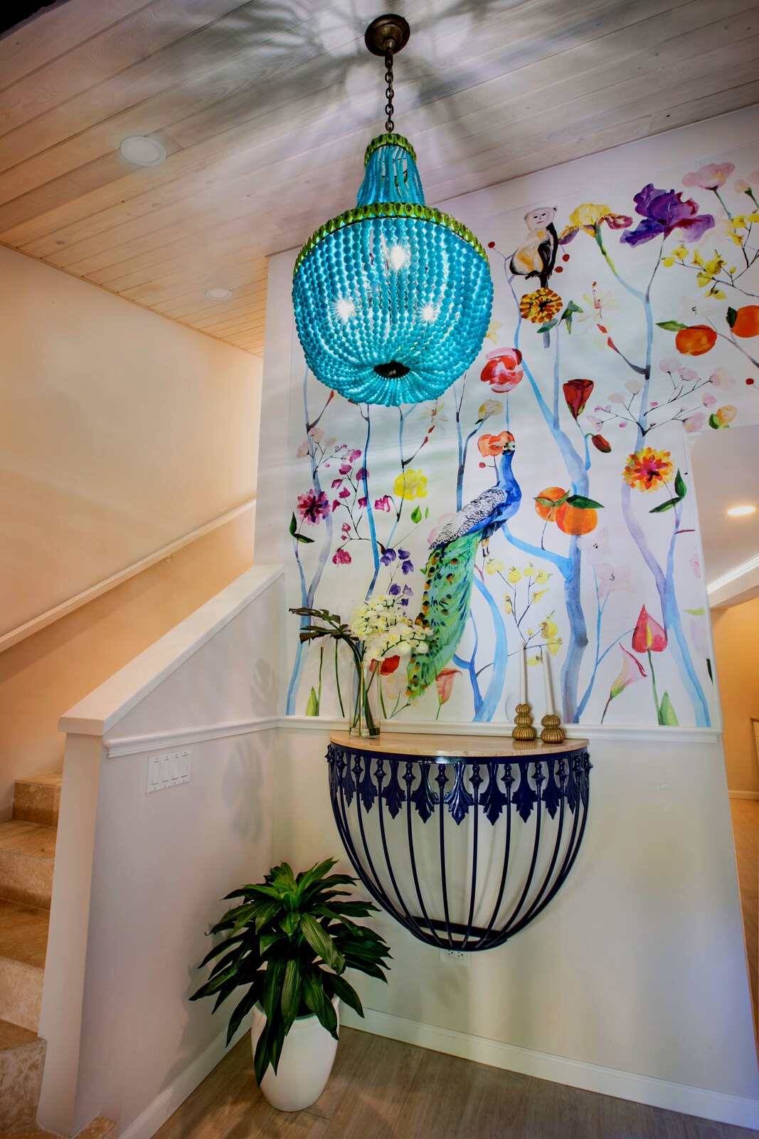 A bright turquoise glass chandelier hangs in a staircase landing with a colorful floral mural on the wall, a black metal console table, and a potted plant below.